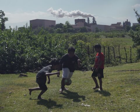 Tiga anak —Syahrul, Rehan, dan Baim— bermain bola di tanah lapang. Asap dari cerobong smelter terlihat membumbung tinggi di belakang mereka/Arya Nur Prianugraha untuk Bollo.id