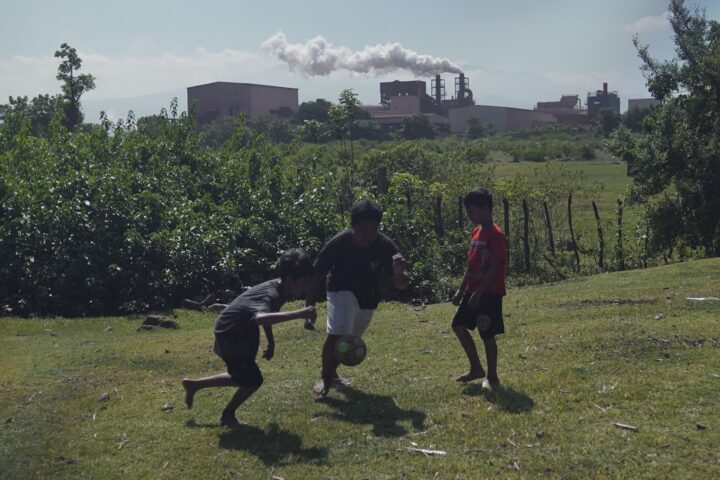 Tiga anak —Syahrul, Rehan, dan Baim— bermain bola di tanah lapang. Asap dari cerobong smelter terlihat membumbung tinggi di belakang mereka/Arya Nur Prianugraha untuk Bollo.id