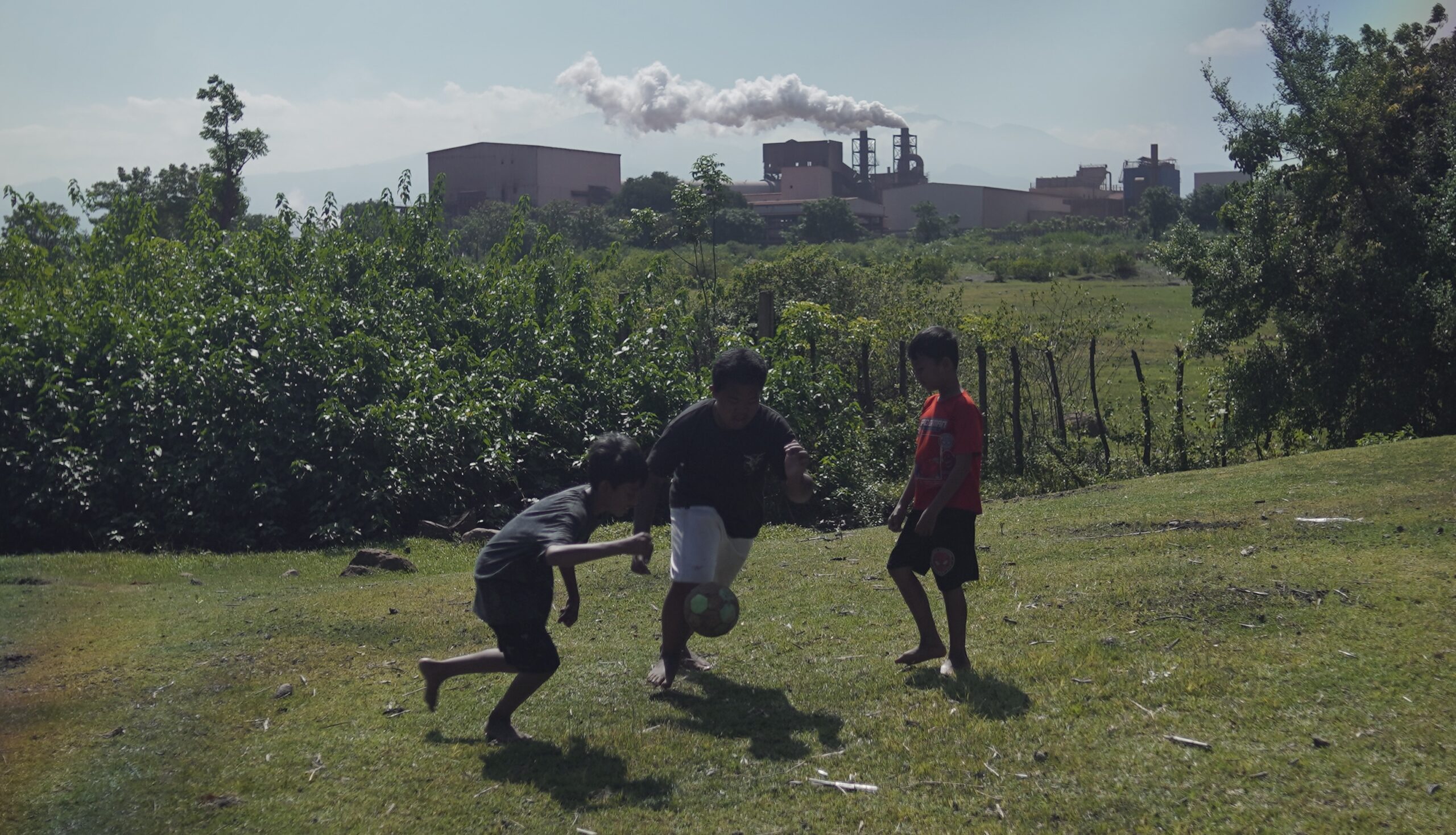 Tiga anak —Syahrul, Rehan, dan Baim— bermain bola di tanah lapang. Asap dari cerobong smelter terlihat membumbung tinggi di belakang mereka/Arya Nur Prianugraha untuk Bollo.id